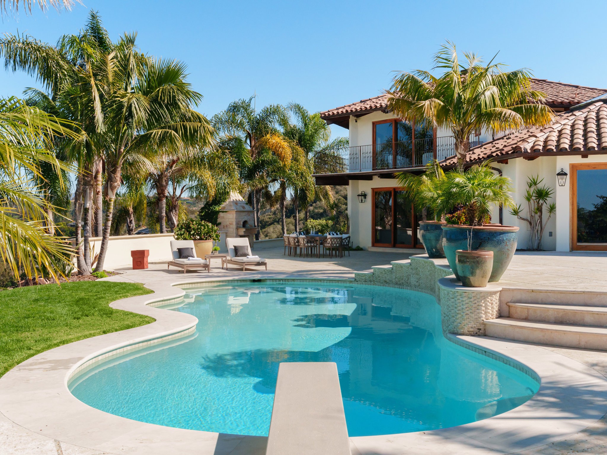 Coronado home's luxurious backyard pool area with sun-kissed patio and lounge chairs surrounded by lush palm trees.