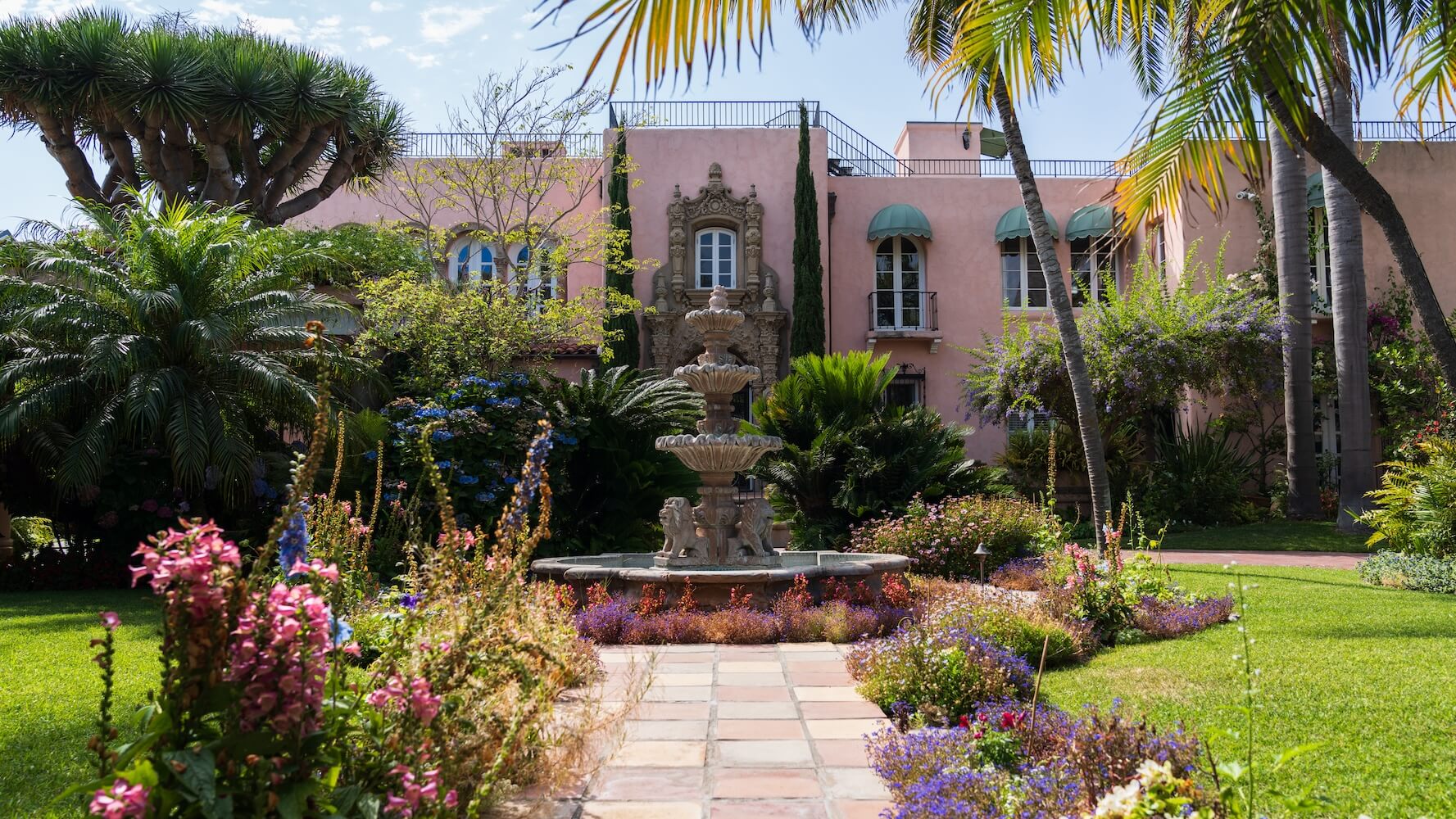 Mediterranean-style luxury mansion in Coronado Village surrounded by lush, manicured landscaping and gardens. A red tile roof contrasts against the clear blue sky.